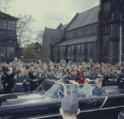 The Queen and the Duke of Edinburgh are being driven in a black convertible. A large crowd of people wave to them. A church stands in the background.