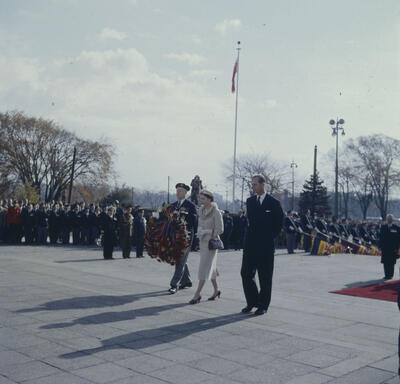 The Queen and the Duke of Edinburgh are accompanied by a man in uniform carrying a large wreath. A crowd of people, many in uniform, stand behind them. A flagpole bears the Canadian flag.