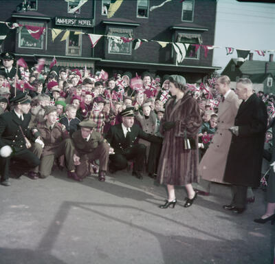 Then-Princess Elizabeth, wearing a long, fur coat, walks past a crowd of people. Several photographers are kneeling before her to take photos.