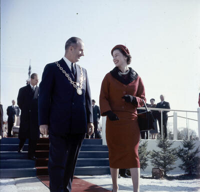 The Queen, in a red dress and matching hat, walks beside and smiles at the Mayor of Ottawa. The Mayor wears a dark suit and the chain of office. 