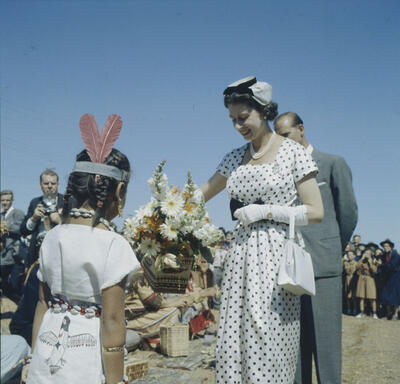 La Reine reçoit un bouquet de fleurs des mains d’une jeune fille autochtone en habits traditionnels. Le duc d’Édimbourg est derrière elle. On voit une foule à l’arrière-plan. 