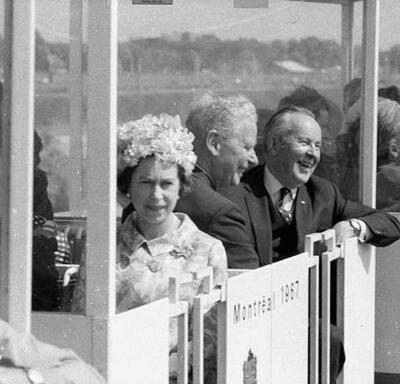 A black-and-white photo of The Queen, in a floral hat, sitting in a trolley, with Lester B. Pearson and another man in the seats behind her. The words “Montréal 1967” are printed on the side of the trolley.