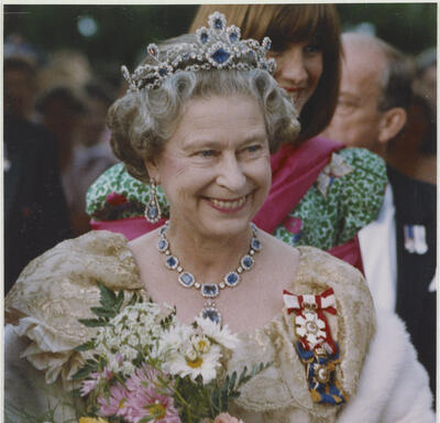 The Queen, holding a bouquet of flowers in her arms, smiles and poses. She is dressed in formal attire and wears the Order of Canada and other insignia. 