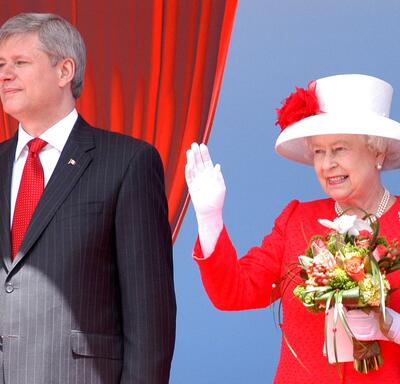 The Queen, in a red outfit and white-and-red hat, holds a bouquet of flowers and waves. She is smiling. Then-Prime Minister Stephen Harper stands next to her, wearing a dark suit with a red tie.