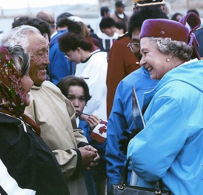The Queen, in a blue parka and burgundy hat, smiles at two Inuit Elders in traditional attire. A child holding a small Canadian flag looks on. A crowd of people are in the background. 