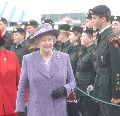The Queen, wearing a purple coat and matching hat, walks past a group of cadets in uniform, standing at attention.