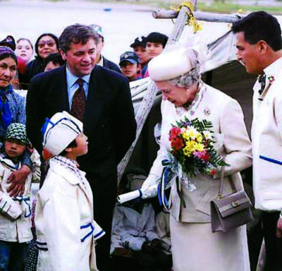The Queen, dressed in white and carrying a bouquet of flowers, is greeted by a child wearing a white-and-blue ceremonial coat and hat. Two officials look on, while a crowd of people stand behind them. 