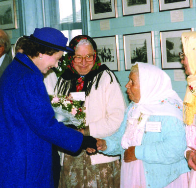 The Queen shakes hands with a woman wearing a kerchief, while two other women in similar attire look on.