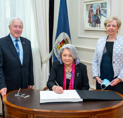 La gouverneure générale Mary Simon est assise à un bureau. Elle a un stylo à la main et signe un livre d'or. Un homme est debout à sa gauche et une femme est debout à sa droite. Ils sont tous souriants.