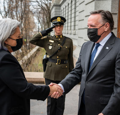 Governor General Mary Simon is shaking hands with the Premier of Quebec. They are outdoors and they are both wearing masks. A guard is standing at attention in the near distance. 