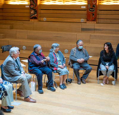 A large group of people is sitting in a circle. They are in a large room with a wooden floor and walls.