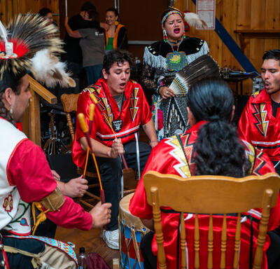 A group of 5 people singing and beating a drum. There are people behind them. They are indoors.