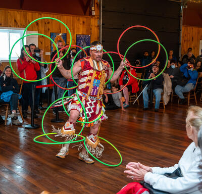 A man is performing a dance while handling several hula hoops. There is a crowd around him. They are indoors.