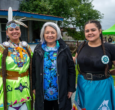 Governor General Simon is standing between two women at Mādahòkì Farm. They are outside and smiling.