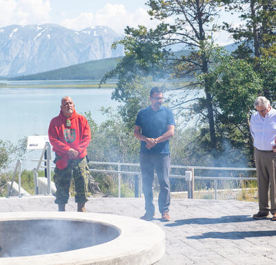 La gouverneure générale Simon, M. Fraser et deux hommes sont debout autour d'une grande fosse à feu en pierre. De la fumée s'élève de la fosse. Derrière eux, il y a un lac et une montagne.