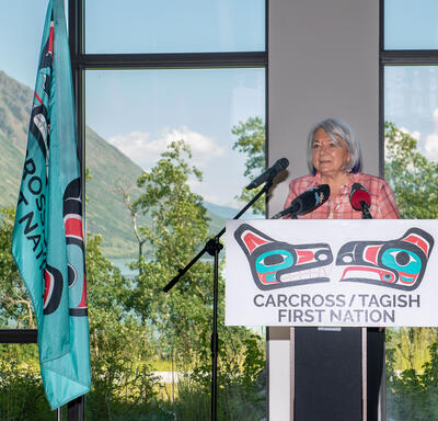 La gouverneure générale Simon se tient sur une plateforme surélevée. Elle parle dans plusieurs microphones. Une affiche devant elle indique : « Première Nation de Carcross/Tagish » (en anglais). Une grande fenêtre derrière elle offre une vue sur une monta