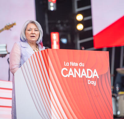 La gouverneure générale est debout derrière un podium ou est inscrit en blanc sur fond rouge “La fête du Canada Day.”