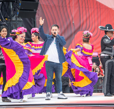 Un artiste chante entouré de danseuses portant des costumes de couleurs pourpre, rose et jaune éclatants sur scène lors de la cérémonie de la fête du Canada.