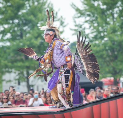 An Indigenous performer dances on stage wearing a mauve costume with feathers at the Canada Day daytime ceremony.