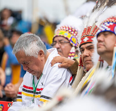 People wearing traditional Indigenous headdresses. A man is bent over with his eyes closed. A man has his hand on his shoulder.