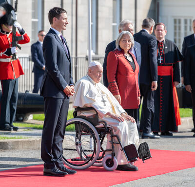 Governor General Simon and Prime Minister Justin Trudeau are standing on either side of Pope Francis who is sitting in a wheelchair. They are on a red carpet.