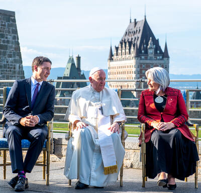 Governor General Simon, Prime Minister Justin Trudeau and His Holiness Pope Francis are sitting next to each other outside of the GGCitadelle. The Chateau Frontenac is in the distance behind them.