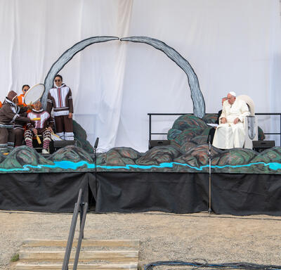 A large stage decorated with mountainous scenery. Performers are singing and playing instruments on the right side of the stage. Pope Francis and another man sit on the left side of the stage. 