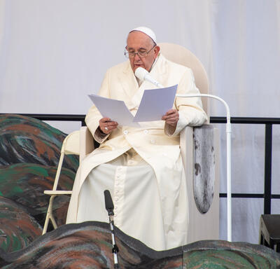 Pope Francis is delivering a speech. He is sitting on a white chair and has sheets of paper in his hands. 