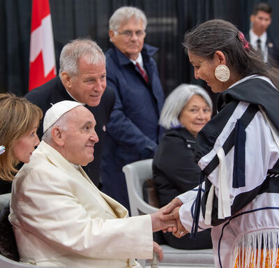 Pope Francis shakes the hand of a woman. The woman is wearing traditional Inuit clothing. Governor General Simon and Mr. Fraser smile in the background.