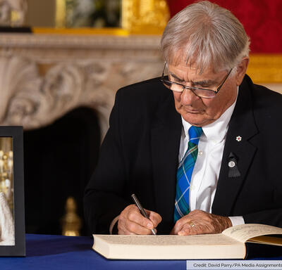 Son Excellence est assise à une table et il écrit dans un livre. Il y a un portrait de Sa Majesté la Reine sur la table. Dans le coin inférieur droit de la photo est écrit : « Source : David Parry/PA Media Assignments ».