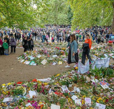 Une foule nombreuse est rassemblée dans un parc. De grandes quantités de fleurs et de cartes posées sur le sol tracent un chemin à travers le parc.