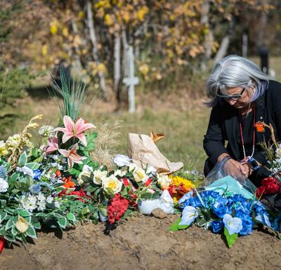 Governor General Mary Simon is kneeling beside a grave covered with flowers. 