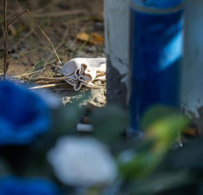 A small tobacco pouch is at the base of a cross. A blue candle and flowers are blurred in the foreground. 