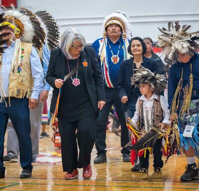 The Governor General is standing in the gymnasium. She is holding a feather, looking down at a young child to her left.