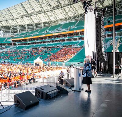 La gouverneure générale Simon est sur une scène dans un stade. Elle se tient derrière un pupitre et parle dans un microphone. Une foule de personnes portant des chandails orange est assise devant la scène.