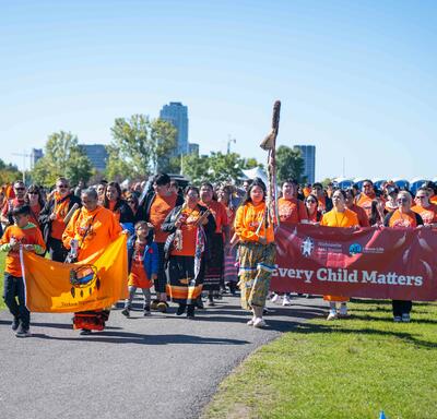 Un groupe de personnes défilant. Beaucoup d'entre eux portent des chandails orange et une grande bannière indiquant "Every Child Matters" est visible.