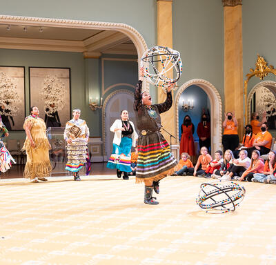 Une danseuse de cerceaux se produisant dans la salle de bal de Rideau Hall. Elle porte une jupe traditionnelle à rubans et tient cinq cerceaux.