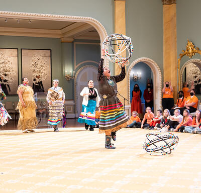 A hoop dancer performing in the Ballroom at Rideau Hall. She is wearing a traditional ribbon skirt and is holding 5 hoops.