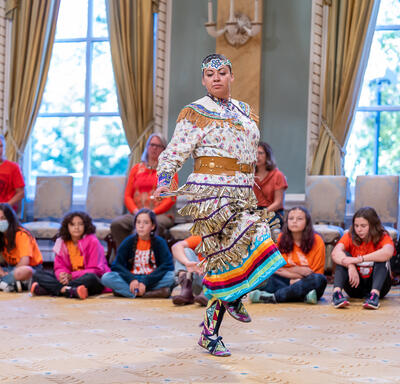 A dancer performing in the Ballroom at Rideau Hall. She is wearing a traditional outfit. Schoolchildren are seated around her, watching.