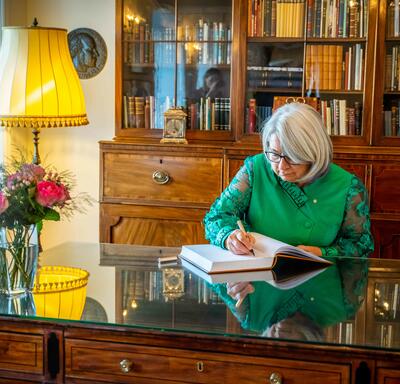 Governor General Mary Simon is sitting at a desk in a study. She is writing in a book. 