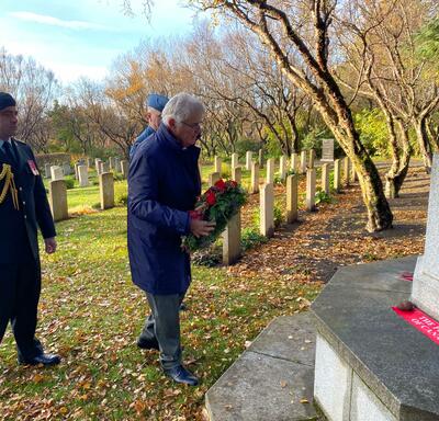 Mr. Fraser, Col. Ronald Walker and a person wearing a military uniform are standing in front of a stone memorial in Fossvogur Cemetery. There is a wreath leaning against the memorial. 