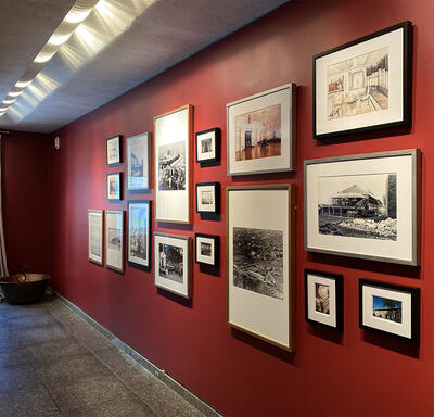 Angled view from the right of a red wall that features a mix of 17 photographs, sketches and paintings on a red wall.