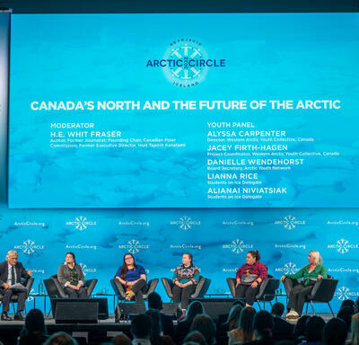 Mr. Fraser and five women are sitting in black armchairs on a stage. A crowd is seated in front of the stage.
