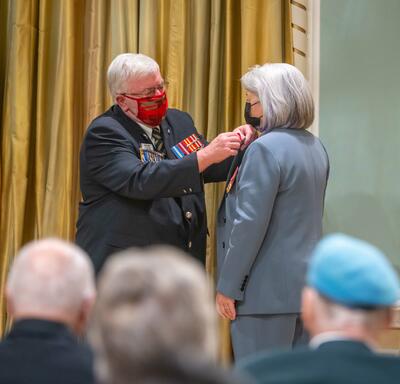 Mr. Bruce Julian, Dominion President of the Royal Canadian Legion pins a poppy to Governor General Simon’s shirt. An audience is seated in front of them.