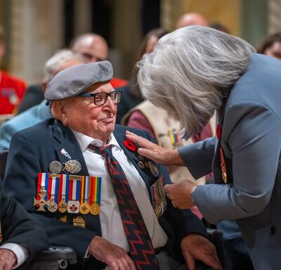 Governor General Simon stands in front of a man who is seated in a wheelchair. She touches a poppy pinned to his shirt. There is a group of people seated around them.