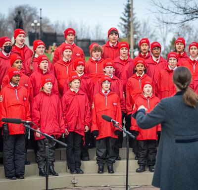 A group of children wearing red coats and hats is singing. They are outside. Someone who has their back to the camera is leading the choir.