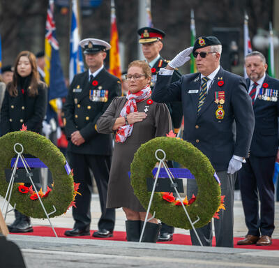 Mme Candy Greff est debout devant deux couronnes de fleurs au Monument commémoratif de guerre du Canada à Ottawa. Un homme est debout à côté d'elle et salue. Plusieurs personnes sont debout derrière eux.
