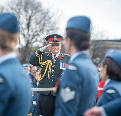 La gouverneure générale Mary Simon salue. Elle porte l’uniforme de l’Armée canadienne. Il y a une foule derrière elle. Des membres des Forces armées canadiennes marchent devant elle et la regardent.