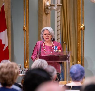 Governor General Mary Simon is wearing a pink suit. She is standing behind a podium in front of a large crowd in the Ballroom of Rideau Hall. A large mirror and a flag of Canada are visible behind her.