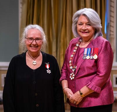 La gouverneure générale Simon se tient à côté d’une femme qui porte une médaille épinglée à son habit noir.